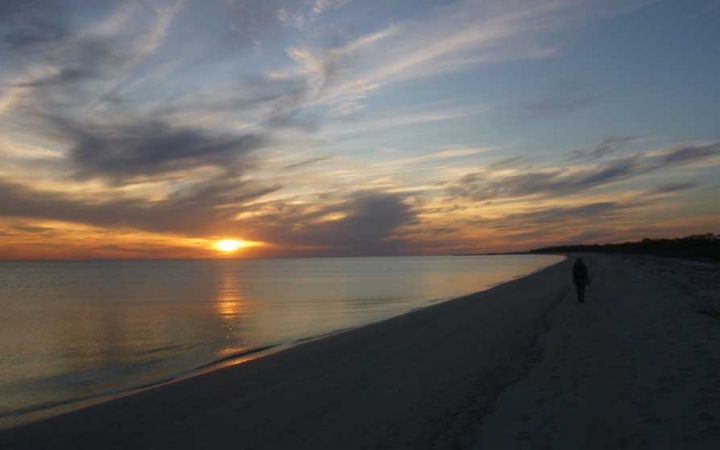 A person walks along a sandy beach next to calm water. The sun is rising or setting along the horizon.